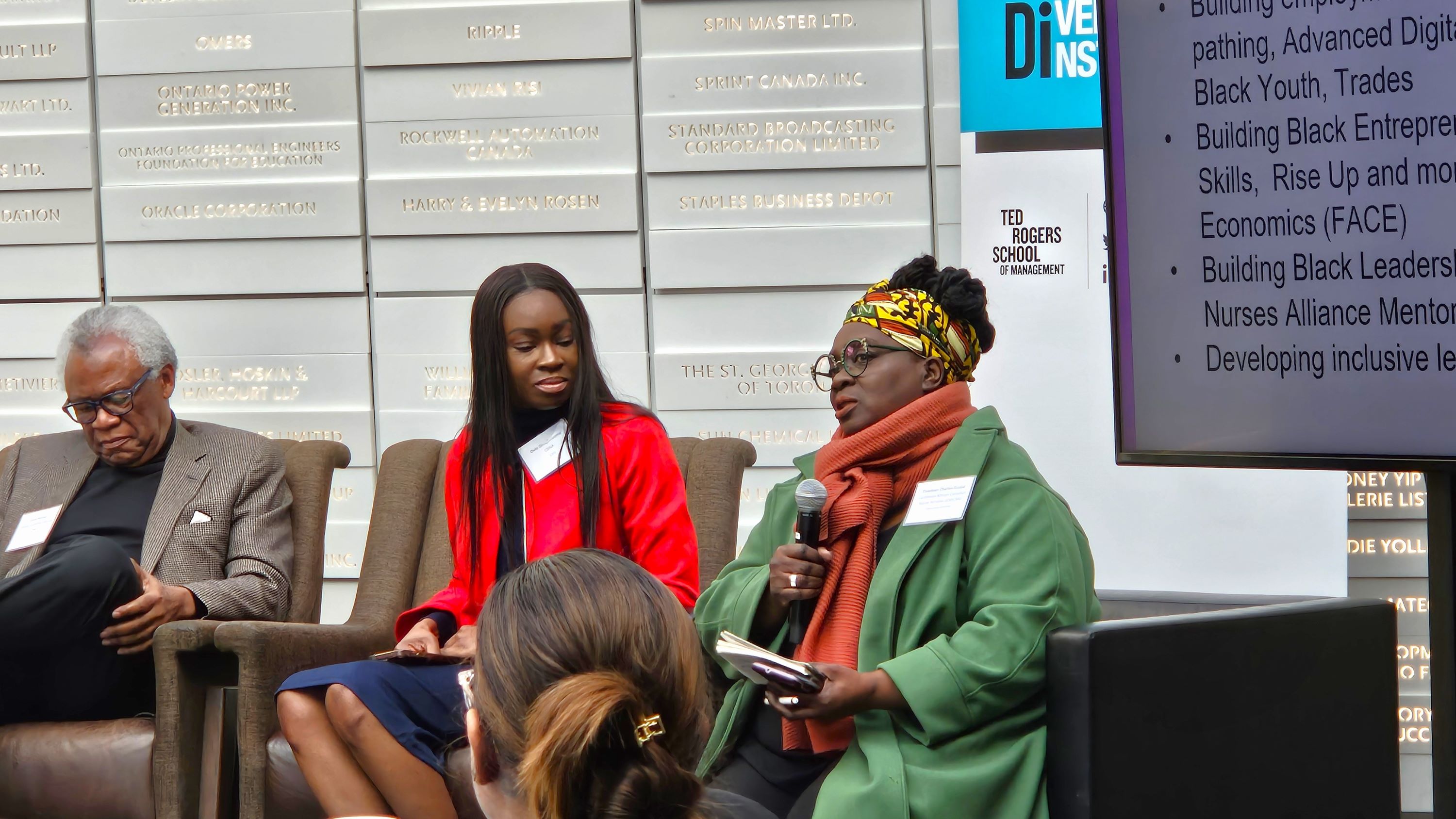 Three Black panelists, one man and two women, sitting on stage, with one of the women speaking into a microphone.