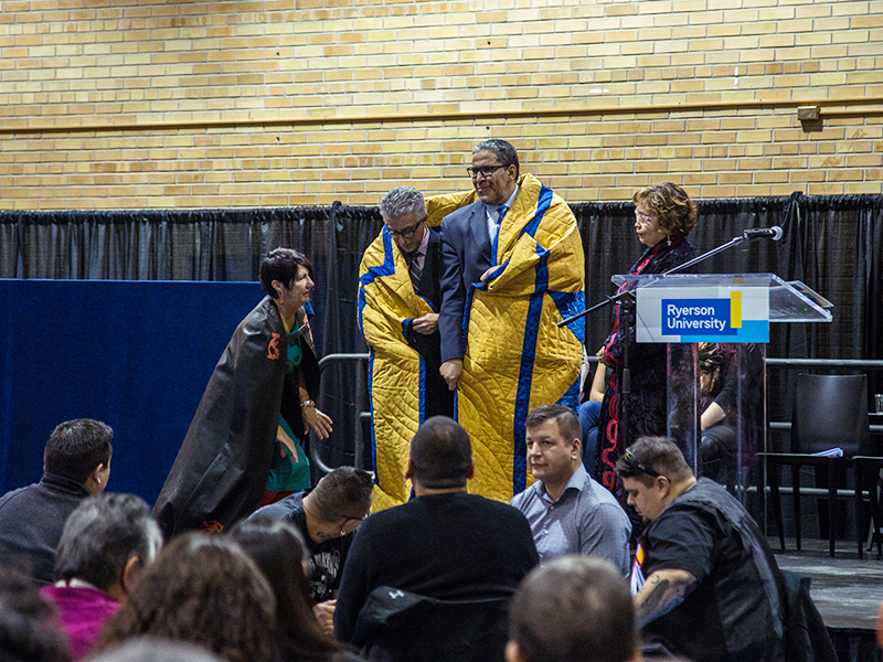 Michael Benarroch and Mohamed Lachemi being wrapped in the yellow and blue star blanket at the Truth and Reconciliation at TMU community celebration